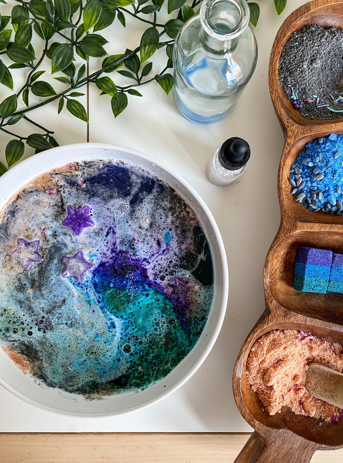 The Magic Folks Sensory Potion Kit in use — a swirling galaxy-themed bath activation with purple star melts, teal and indigo color-changing water, and blue, black, and pink sensory sand accents arranged in a wooden dish