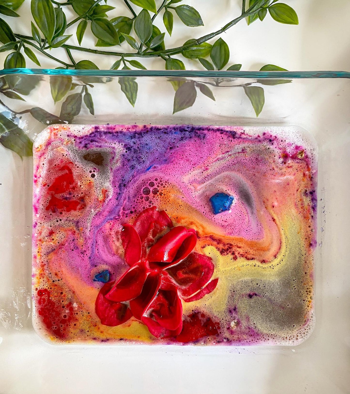 Close-up of a sensory potion kit color-changing bath water swirling in pink, purple, orange, and yellow, with a red rose in bloom and a blue gem-shaped melt floating on the surface