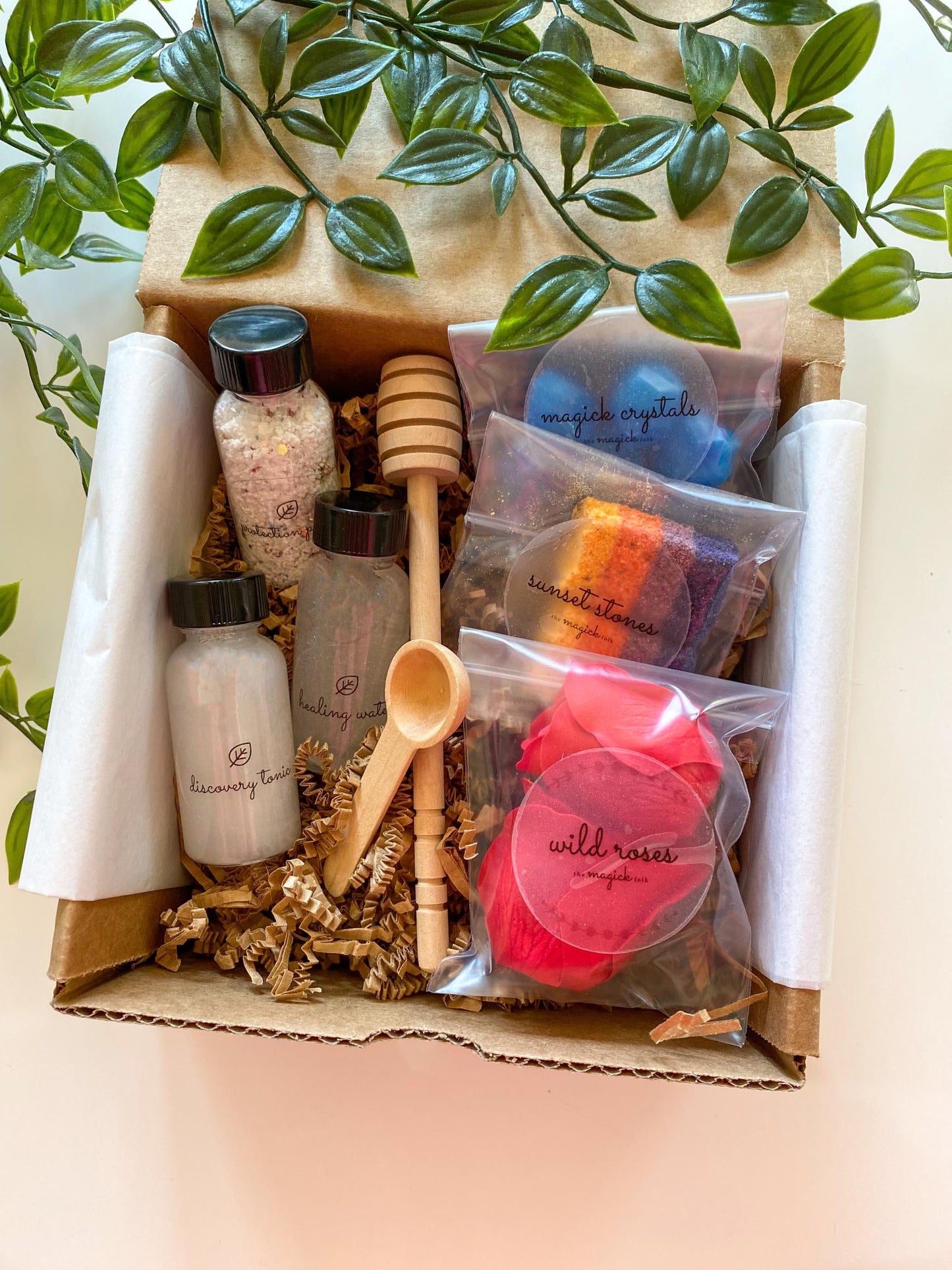 Unboxed sensory potion kit with labeled bottles and jars, sealed bags of blue crystal-shaped pieces, striped sunset-colored bath chunks, and red rose petals, packaged in a kraft box with a wooden scoop and dipper